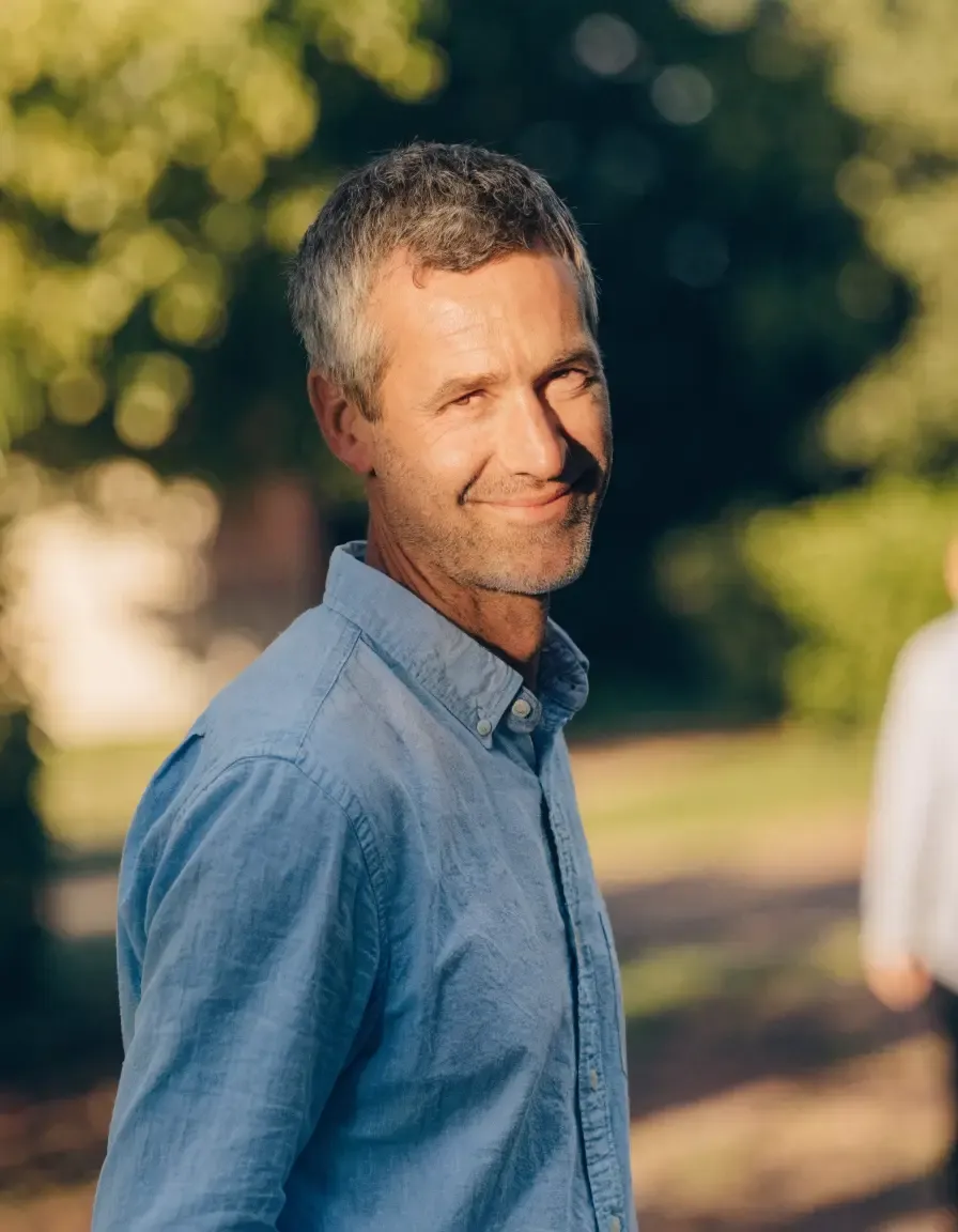 Middle-aged man with gray hair in a blue shirt smiling gently outdoors.